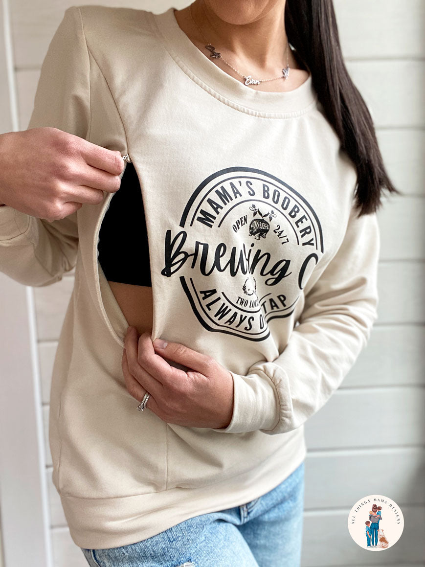 Women wearing a beige crewneck sweatshirt with a design logo Mama's Boobery Brewing Co Open 24/7 Always on Tap Two Locations in black with two zippers on either side for easy access to breastfeed or pump. Right side of zipper is open to show how it works.