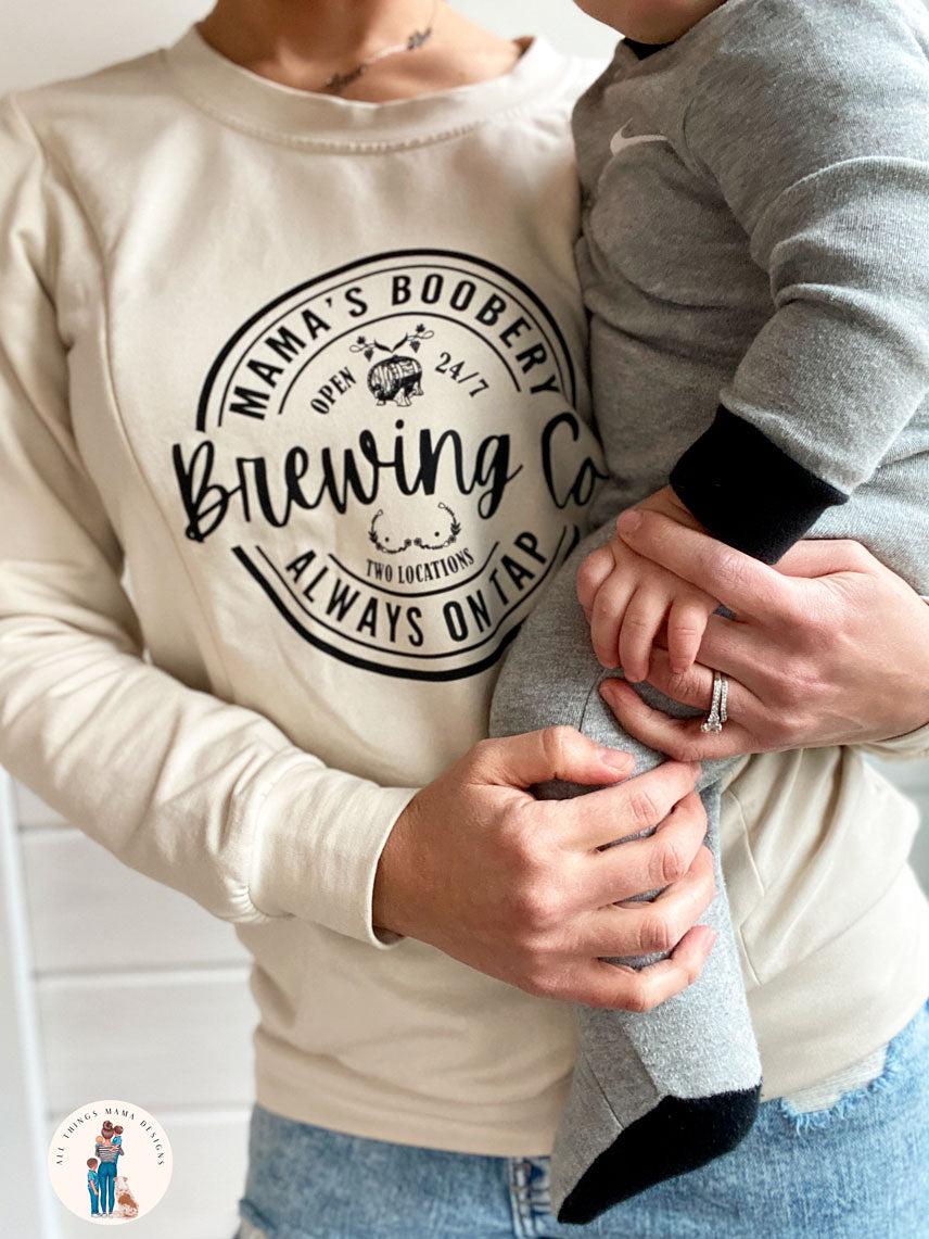 Women wearing a beige crewneck sweatshirt with a design logo Mama's Boobery Brewing Co Open 24/7 Always on Tap Two Locations in black with two zippers on either side for easy access to breastfeed or pump and holding a small baby.