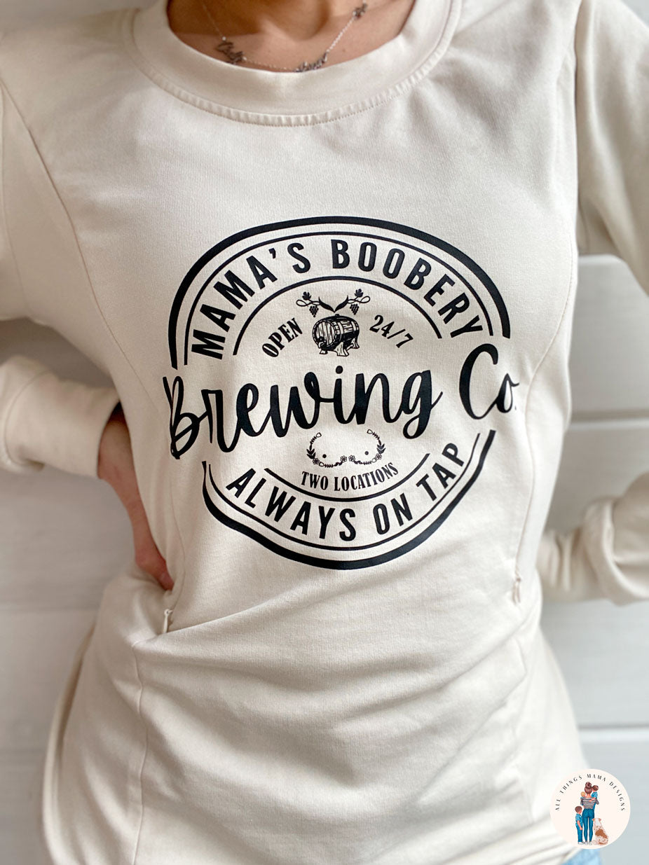 Women wearing a beige crewneck sweatshirt with a design logo Mama's Boobery Brewing Co Open 24/7 Always on Tap Two Locations in black with two zippers on either side for easy access to breastfeed or pump. Right side of zipper is open to show how it works. Sweatshirt is breastfeeding friendly