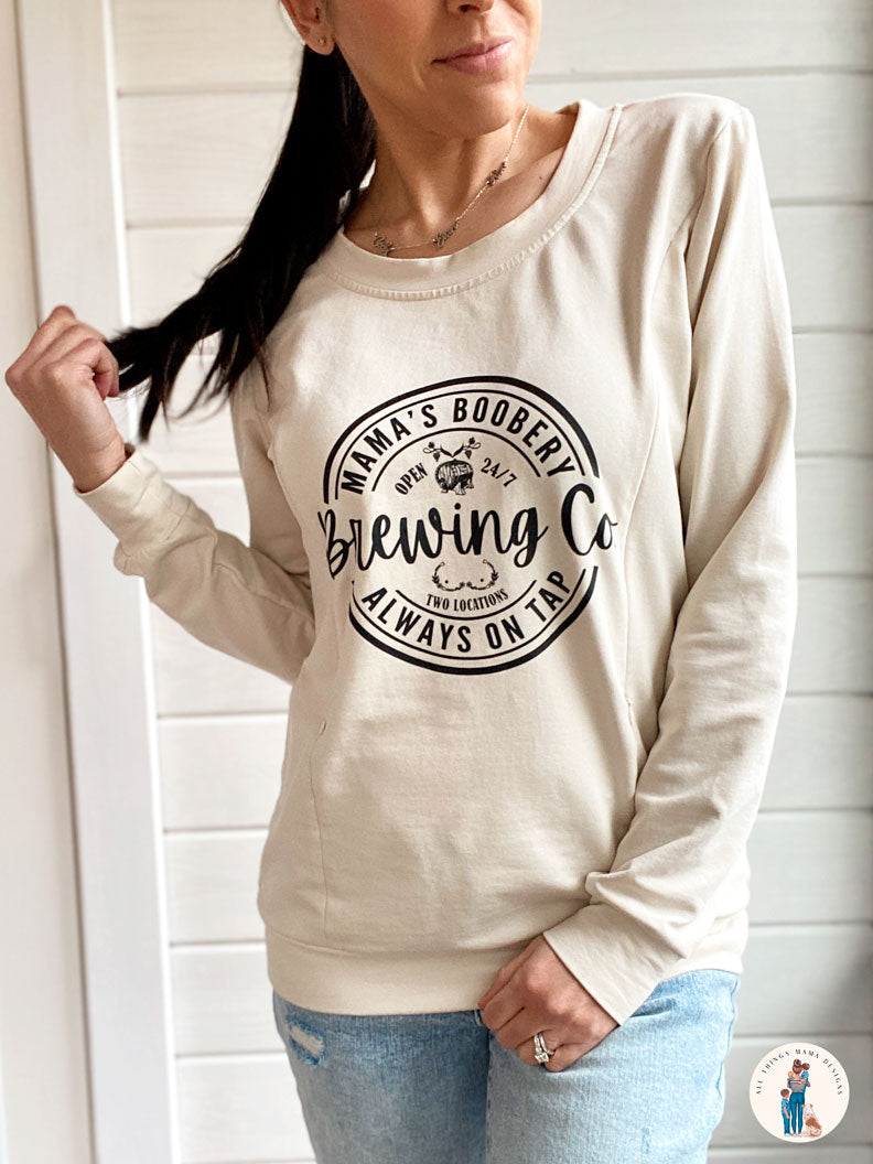 Women wearing a beige crewneck sweatshirt with a design logo Mama's Boobery Brewing Co Open 24/7 Always on Tap Two Locations in black with two zippers on either side for easy access to breastfeed or pump