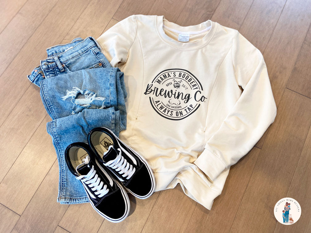 Beige crewneck sweatshirt with a design logo Mama's Boobery Brewing Co Open 24/7 Always on Tap Two Locations in black with two zippers on either side for easy access to breastfeed or pump. Displayed with a ripped pair of jeans and black and white Vans sneakers.